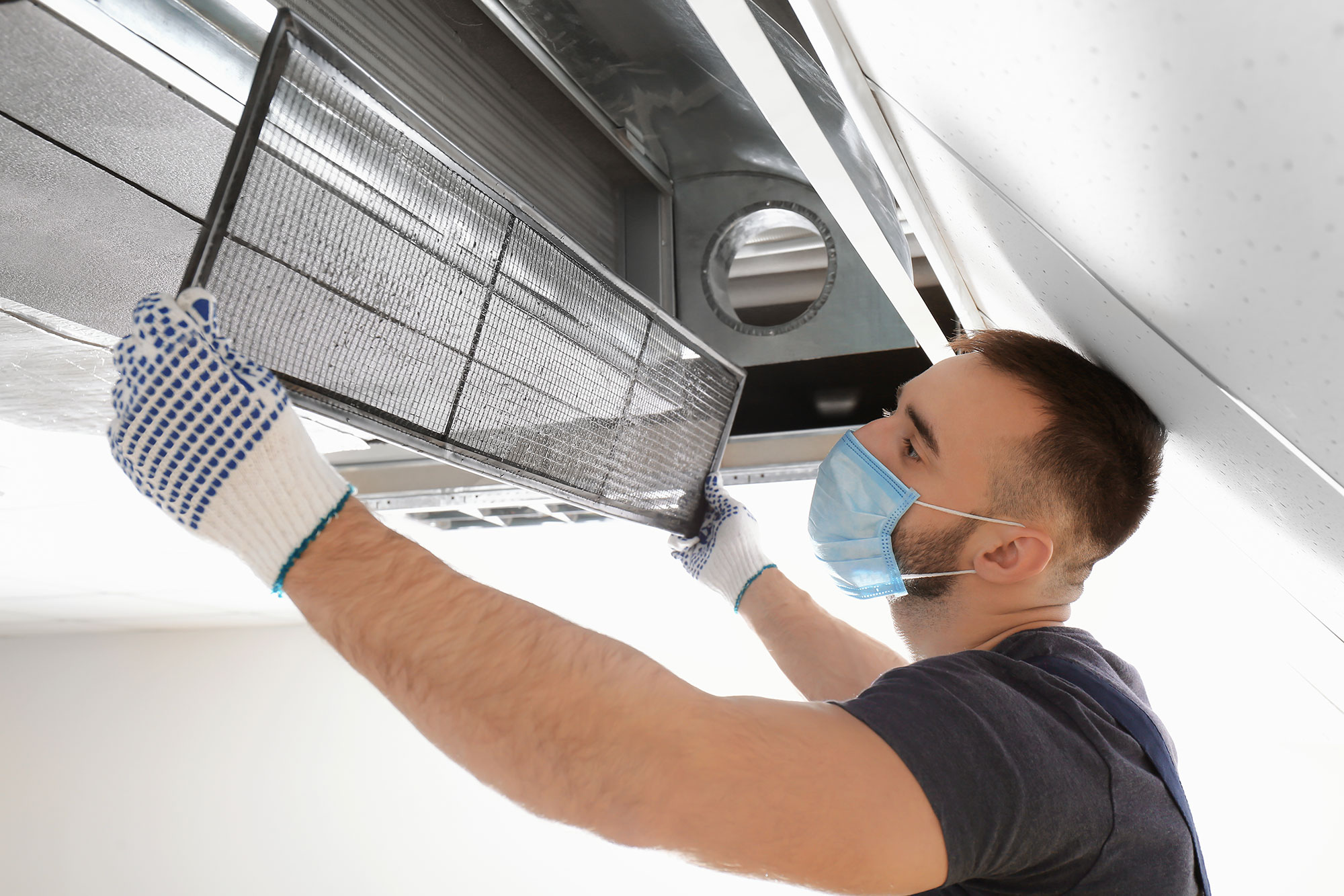 Technician wearing gloves and a face mask removing a mesh air filter from a ceiling mounted ventilation unit