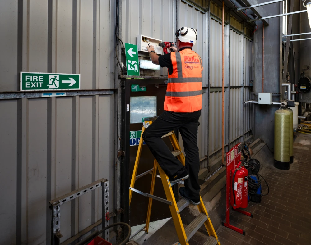 Engineer installing emergency lighting above a signed fire exit route in an industrial unit