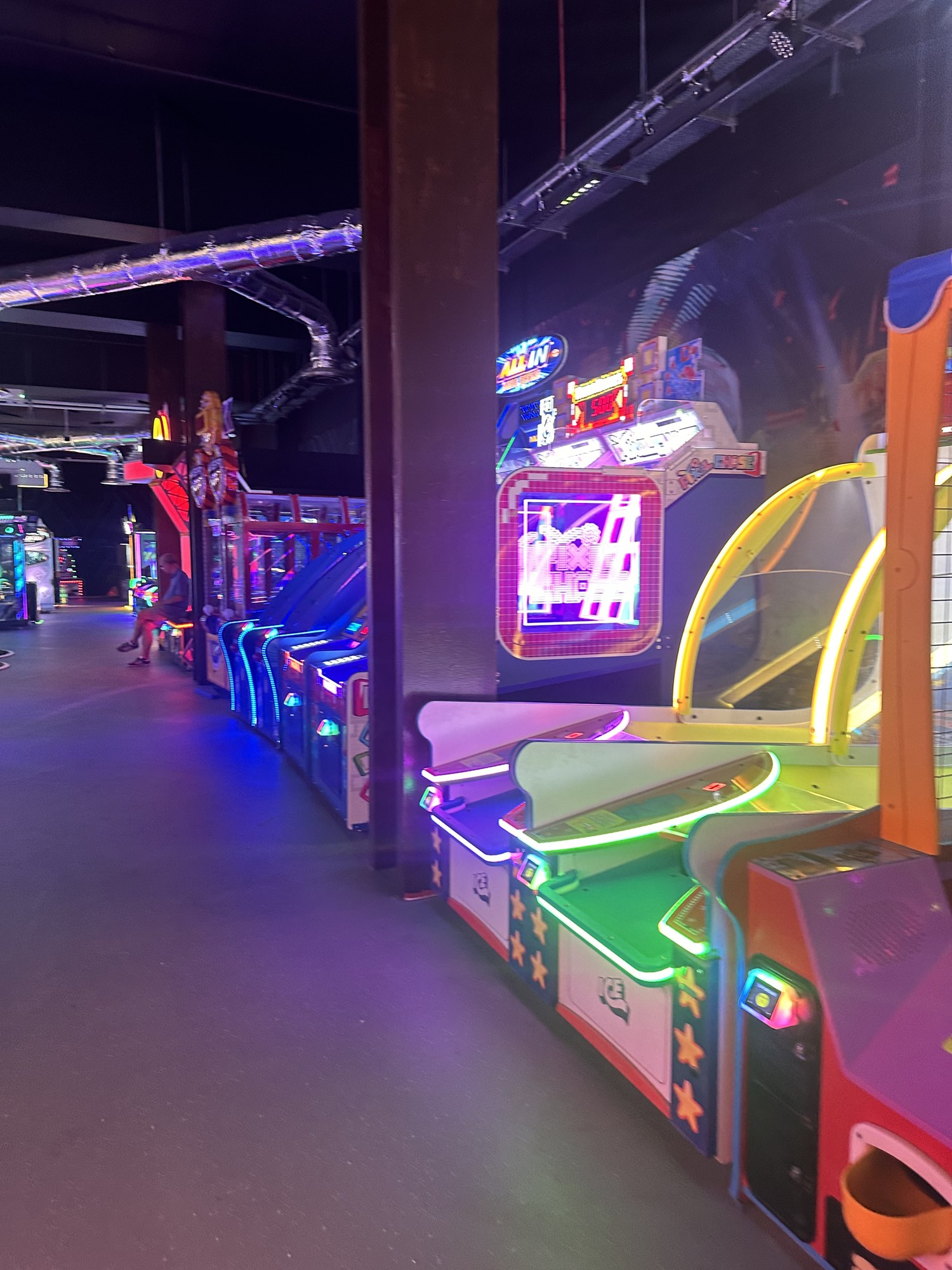 Neon lit arcade machine lines with illuminated signage and exposed spiral ductwork above